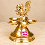 Side profile view of a traditional Brass Panchmukhi Peacock Design Diya displayed against a clean premium beige studio background, highlighting the sculpted golden brass peacock finial, five surrounding oil lamp bowls, decorative supporting column, and broad pedestal base. The side angle reveals the peacock's gracefully curved neck, detailed head, textured feather patterns, and elaborately raised tail, showing the depth and dimension of the ornamental bird figure. The shallow brass diya bowls extend outward