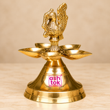 Three-quarter angled view of a traditional Brass Panchmukhi Peacock Design Diya featuring an intricately carved golden brass peacock figure positioned above five decorative oil lamp bowls arranged around a central supporting column. The angled perspective highlights the dimensional shape of the peacock, including its gracefully curved neck, detailed feather carvings, decorative wing patterns, and raised tail with ornamental texturing. The five shallow brass diya bowls form a symmetrical circular arrangement