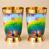 Pair of brass water glasses with colorful peacock printed design, polished golden interior, decorative gold-tone rim and sturdy base, presented on a premium beige studio background for elegant serving and special occasions.