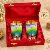 Pack of 2 peacock printed brass water glasses presented in a red gift box, featuring polished golden interiors, colorful peacock artwork and decorative gold-tone rims and bases, suitable for festive and special occasion gifting.