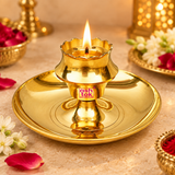 Traditional polished brass diya shown in use in a warm devotional setting with a gently burning flame in the raised central holder, surrounded by tasteful flowers, rose petals and subtle brass puja accessories on a beige-toned surface, demonstrating practical use for daily worship, evening aarti, Diwali lighting, festive puja, religious ceremonies and home mandir rituals without any deity idol included in the scene.