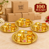 Brass puja thali set bulk display for 100 piece set with multiple thali sets arranged in front of plain packaging boxes on a beige background.