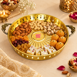 Round brass serving tray styled in a festive lifestyle setting and arranged with almonds, cashews, pistachios, raisins, Indian sweets and laddus to demonstrate practical serving use. The polished golden tray features ornate floral handles on both sides, a decorative perforated rim and an embossed radial petal design across the base. Surrounded by subtle festive décor, flowers and traditional accents on a warm beige setting