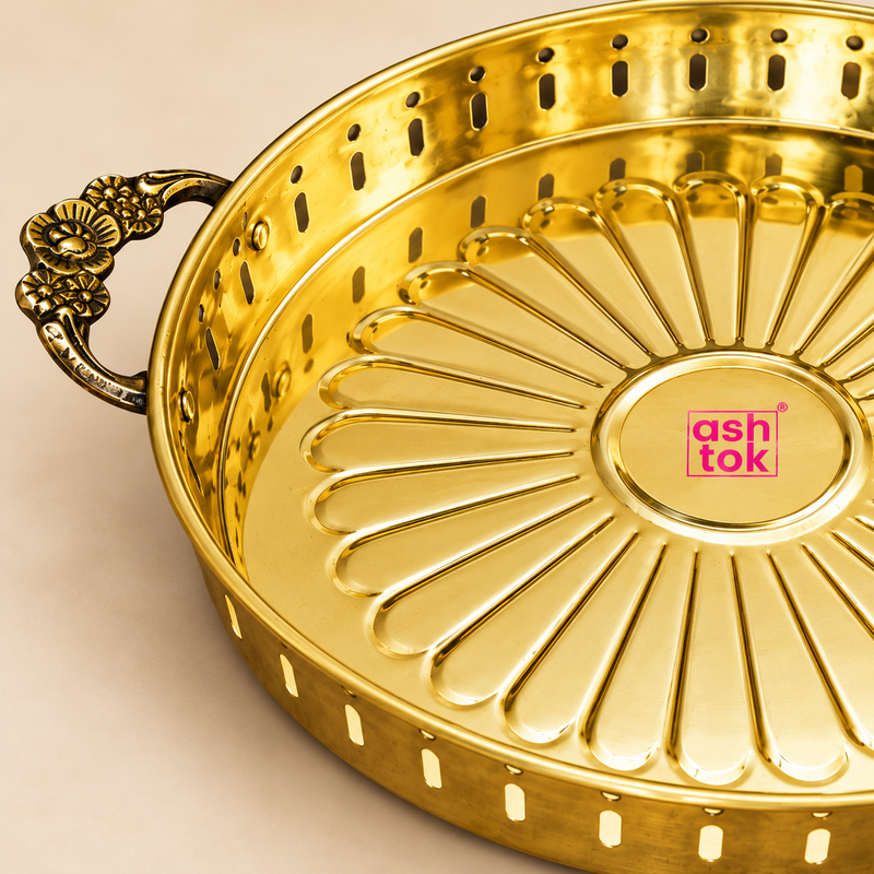Detailed close-up view of a polished golden brass serving tray highlighting the ornate floral side handle, decorative perforated side wall and embossed flower-petal pattern on the serving base. The close framing shows the craftsmanship of the antique-style handle, smooth polished brass surface, rounded rim and raised radial detailing that gives the tray its traditional decorative character. 