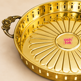 Detailed close-up view of a polished golden brass serving tray highlighting the ornate floral side handle, decorative perforated side wall and embossed flower-petal pattern on the serving base. The close framing shows the craftsmanship of the antique-style handle, smooth polished brass surface, rounded rim and raised radial detailing that gives the tray its traditional decorative character. 
