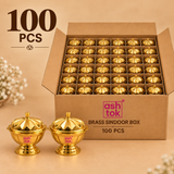 Bulk quantity image of 100 polished brass sindoor boxes arranged neatly inside a large plain brown corrugated cardboard carton with individual divider compartments, with two complete brass sindoor boxes displayed in front to clearly show the rounded bowl shape, decorative petal-style lid, small top knob, polished golden brass finish and pedestal base, presented on a warm beige background for wholesale orders, bulk gifting, festive distribution, wedding return gifts, religious functions and commercial supply