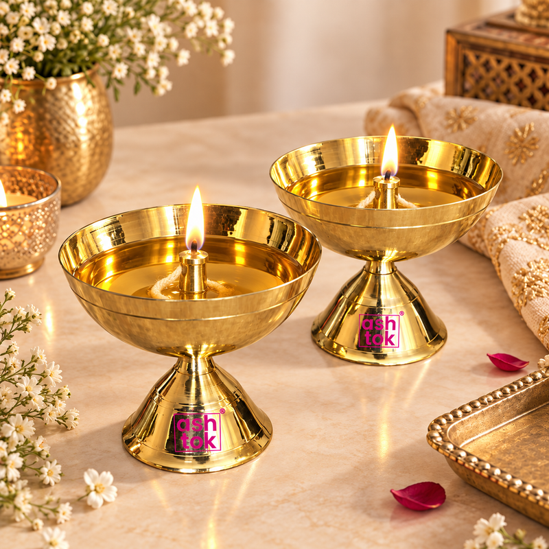 Warm lifestyle photograph featuring two lit polished brass pedestal diyas with burning cotton wicks set on a marble surface, surrounded by white baby's breath flowers, rose petals, a brass serving tray, and traditional festive decor with Ashtok watermarks.