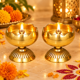 Two Brass Tamatar Diyas illuminated with warm golden flames in an elegant traditional pooja setting, surrounded by fresh marigold flowers, festive decorations, and softly glowing background lights. Each diya features a rounded polished brass bowl, decorative scalloped rim, central wick holder, and beautifully ribbed pedestal base. The glowing brass oil lamps create a devotional atmosphere suitable for Diwali celebrations, daily home mandir prayers, Lakshmi pooja, traditional ceremonies, and festive worship 