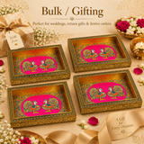 Group display of four identical rectangular Meenakari peacock decorative serving trays arranged together on a festive surface to showcase bulk ordering options, surrounded by fresh white flowers, red rose petals, a wrapped gift box with a silk ribbon, and a glowing brass candle holder, ideal for wedding return gifts and festive hampers.