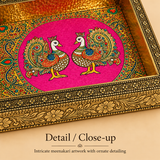 Extreme macro close-up photograph showcasing the intricate Meenakari enamel artwork featuring two detailed peacocks with colorful feathers on a bright pink backdrop, paired with a hammered gold interior wall and an exquisitely carved antique brass floral border.
