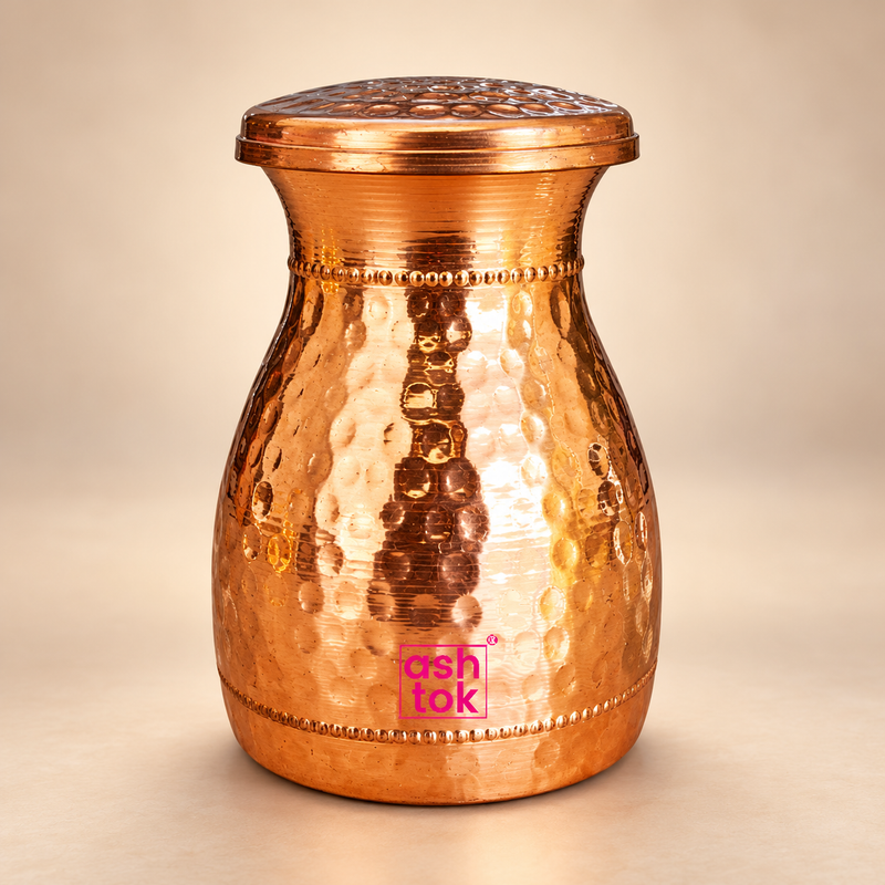 Hammered copper carafe with fitted lid, rounded body, decorative beaded detailing and polished copper finish, presented on a premium beige studio background for water storage and serving.