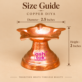 Detailed size guide image of a traditional Copper Diya showing a diameter of 2.5 inches and an overall height of 2 inches, displayed against an elegant beige studio background with clearly marked measurement arrows. The diya features a distinctive flower-shaped upper bowl with multiple outward-facing petals, gently curved pointed edges, and a shallow central oil-holding cavity designed for traditional pooja lighting. Its polished reddish-copper surface displays warm metallic reflections, fine handcrafted-st