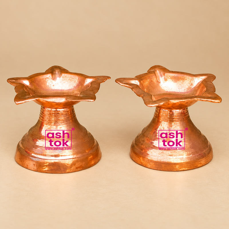 Two traditional Copper Diyas displayed side by side on a premium beige studio background, featuring a beautifully designed flower-shaped oil lamp bowl with multiple curved and pointed petal edges, a polished reddish-copper finish, and a decorative pedestal stand. Each copper diya showcases a distinctive floral design with layered petal-like extensions surrounding the central shallow bowl, creating an attractive traditional appearance suitable for Indian religious rituals. The upper copper bowl features smoo