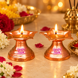 Two traditional flower-shaped Copper Diyas beautifully illuminated with glowing cotton wicks and warm golden flames in an elegant festive pooja setting. Each diya features a polished reddish-copper finish, a shallow oil-holding bowl surrounded by decorative pointed floral petals, and a tapered pedestal stand with a broad circular base. The warm flames reflect across the shiny copper surfaces, highlighting the natural orange-red colour, textured metal finish, and graceful petal-shaped edges of the oil lamps.