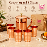 Copper Jug and 6 Glasses drinkware set displayed in an elegant festive gifting arrangement with multiple complete sets, decorative gift boxes, ribbons, flowers and warm beige styling, highlighting the product as a premium option for return gifts, wedding gifts, festive gifting, corporate gifting, housewarming gifts and bulk orders, while showcasing the polished copper jug with lid, curved handle and six matching copper glasses in a coordinated traditional drinkware set with ASHTOK branding.