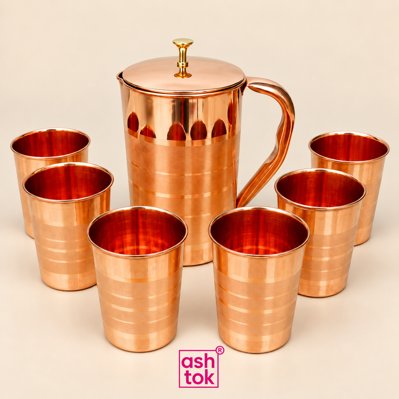 Copper jug and 6 matching copper glasses arranged together as a complete 7-piece drinkware set on a clean beige background, featuring a tall copper jug with fitted lid, decorative knob, smooth curved handle and matching polished copper tumblers with subtle horizontal detailing, presented in a neat ecommerce product composition with ASHTOK branding, ideal for customers looking for traditional copper drinkware for home serving, water storage, guest serving, festive use, wedding gifting, return gifts and premi