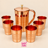 Copper jug and 6 matching copper glasses arranged together as a complete 7-piece drinkware set on a clean beige background, featuring a tall copper jug with fitted lid, decorative knob, smooth curved handle and matching polished copper tumblers with subtle horizontal detailing, presented in a neat ecommerce product composition with ASHTOK branding, ideal for customers looking for traditional copper drinkware for home serving, water storage, guest serving, festive use, wedding gifting, return gifts and premi