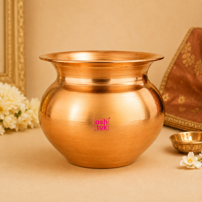 Side profile of a polished copper kalash for pooja decorated with white flowers and a small brass oil lamp.