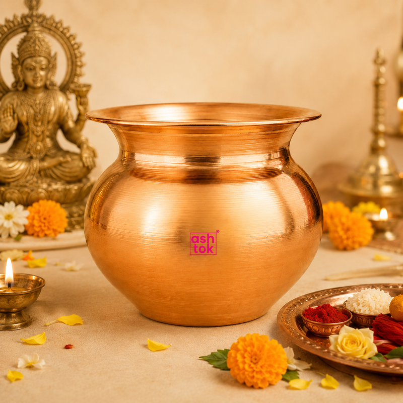Traditional polished copper kalash (handi) placed on a table surrounded by marigold flowers, a brass oil lamp, a statue of Goddess Lakshmi, and a puja thali.