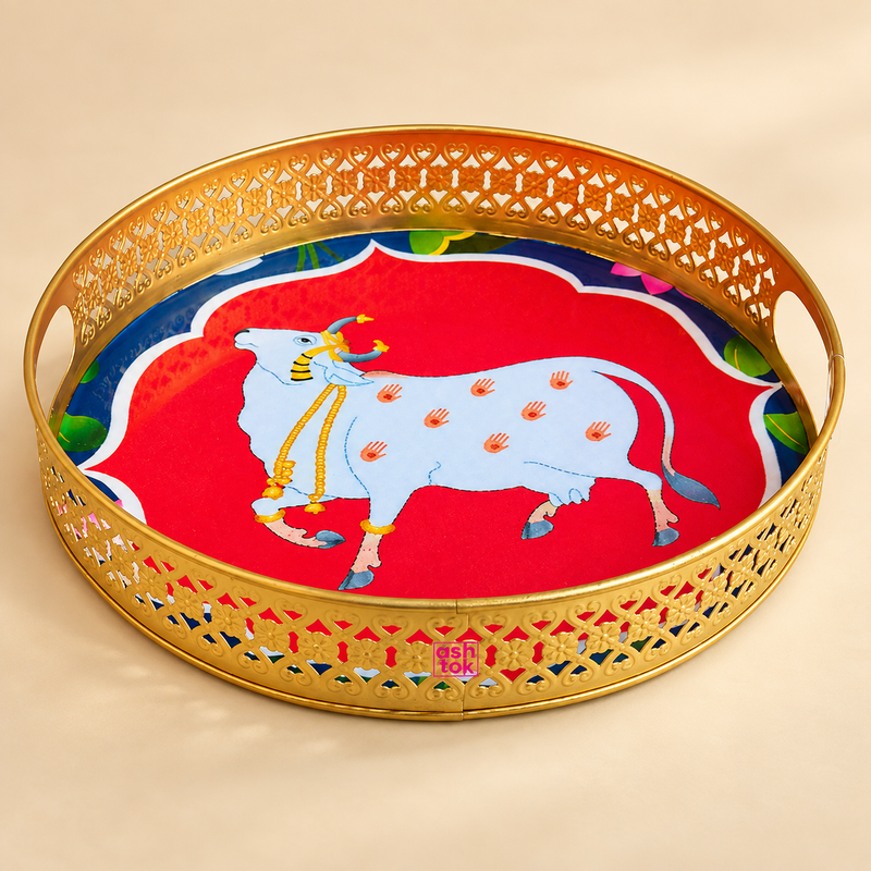 Cow Design Tray Flower Basket and Puja Tray for Temple featuring a round decorative tray with a polished golden brass-look finish, intricate cutwork rim, side carry openings and a vibrant sacred cow artwork in the center. The tray includes the ASHTOK logo on the front and is professionally displayed on a premium beige studio background, making it suitable for puja flowers, prasad serving, festive offerings, temple rituals and traditional home decor use.