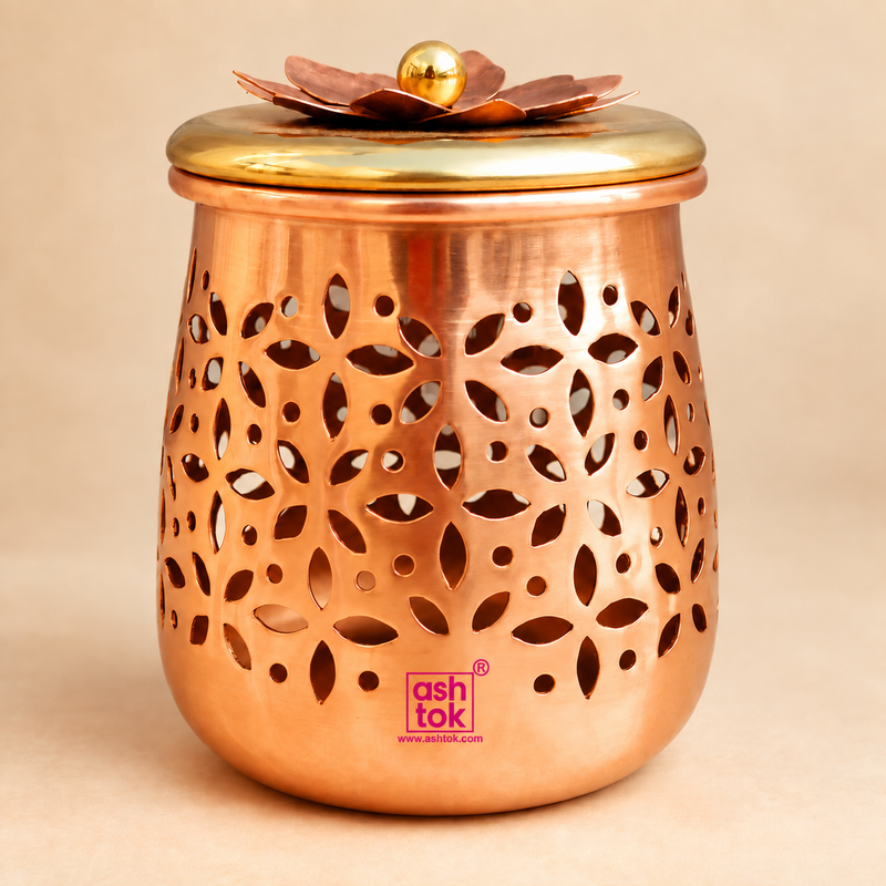 Premium decorative dry fruit gift jar box displayed as a single elegant copper-toned storage jar on a clean warm beige studio background. The jar features a rounded cylindrical body with intricate floral and leaf-shaped cutwork patterns across the exterior, a smooth polished copper finish and a contrasting golden lid decorated with a layered flower design and round top knob.