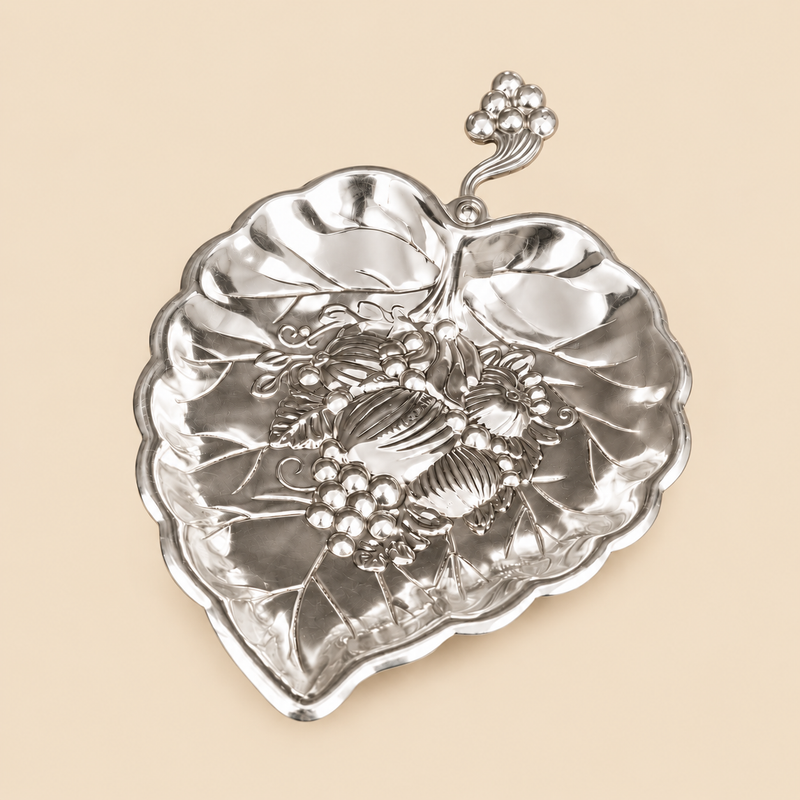 German silver leaf-shaped gift item with embossed floral and fruit design, decorative stem handle and polished silver finish on a beige background.
