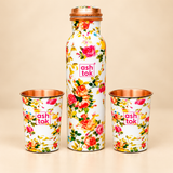 Front view of a floral printed copper water bottle with two matching drinking glasses arranged symmetrically on a beige background. The bottle features a slim cylindrical shape, narrow neck, copper-coloured lid and colourful floral artwork. Both copper glasses have matching rose patterns and visible copper interiors.