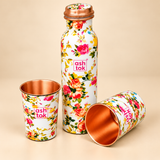 Angled product view of a floral printed copper water bottle with two matching copper glasses, one standing upright and another positioned sideways to reveal its polished copper interior. The white floral exterior displays pink roses, yellow flowers and green leaves, while the bottle features a copper-coloured screw cap.