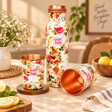 Floral printed copper water bottle with two matching copper drinking glasses arranged on a dining table in a bright home interior. The white bottle and glasses feature colourful rose patterns, polished copper interiors and a copper-coloured lid. Decorative flowers, natural lighting and home accessories demonstrate the set's use for serving drinking water at home.
