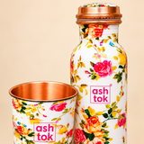 Close-up image of a floral printed copper water bottle beside a matching copper drinking glass, highlighting the pink, red, yellow and orange rose patterns, green foliage, smooth white exterior, copper screw cap and polished copper glass interior. Displayed against a soft beige background to show the decorative finish and product details.