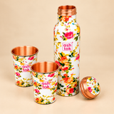 Floral printed copper water bottle shown with its screw cap removed alongside two matching copper drinking glasses. The image highlights the bottle's copper threaded neck, removable copper-coloured cap, polished copper glass interiors and colourful floral printed exterior on a beige studio background.