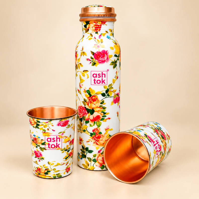 Floral printed copper water bottle with two matching copper drinking glasses displayed on a beige studio background. The set features a white exterior decorated with pink, red, orange and yellow roses, green leaves, a copper-coloured screw cap and polished copper interiors. Suitable for everyday drinking water, home dining, office use and gifting