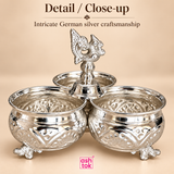 Close-up image of German silver chopala gift item showing the intricate embossed craftsmanship on the bowls, the polished silver finish, the ornamental raised feet and the decorative central finial in sharp detail, presented on a beige background with ASHTOK branding, emphasizing the traditional styling, elegant metalwork, smooth reflective surface and fine detailing that make the piece suitable for gifting, décor and festive utility.