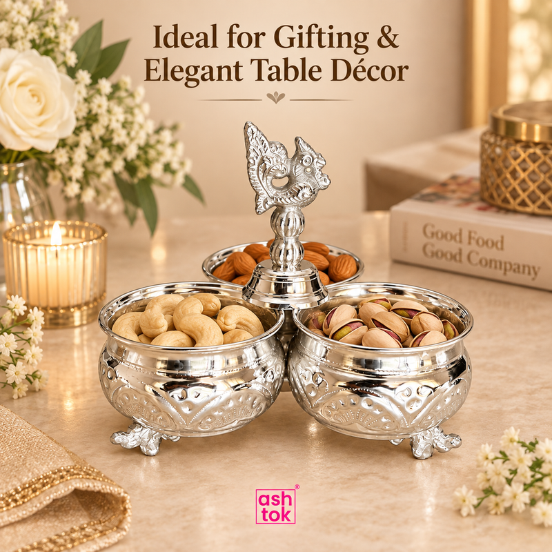 German silver chopala gift item styled in an elegant home setting and used as a decorative serving and display piece, with the three bowls filled with dry fruits such as cashews, almonds and pistachios, placed on a beige tabletop with candles, flowers and books in the background, showing its suitability for festive hosting, return gifting, table décor, special occasions, premium presentation and elegant serving during gatherings and celebrations.
