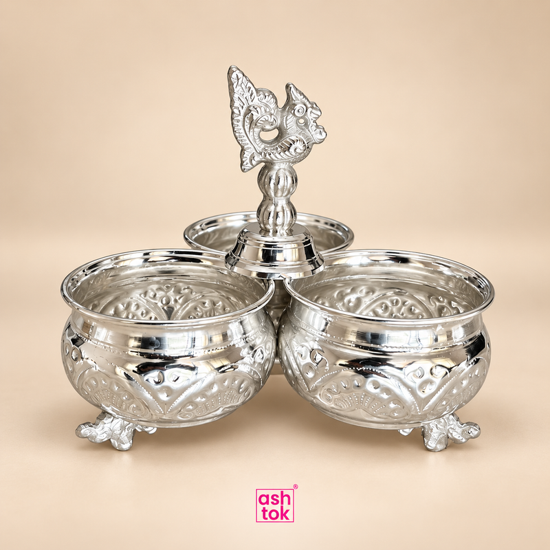 German silver chopala gift item with three round utility bowls joined in a traditional triangular arrangement, featuring embossed decorative detailing, raised ornamental feet and an elegant central finial, displayed as a clean product image on a soft beige background with ASHTOK branding, suitable for gifting, festive presentation, pooja accessory organization, table styling and decorative home use.