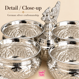 Close-up image of the German silver chopala gift item showing the intricate embossed metalwork on the 4 attached bowls, the highly polished silver surface, the elegant decorative central finial and the ornamental feet in sharp detail, presented on a beige background with ASHTOK branding to emphasize craftsmanship, shine, texture, traditional styling and premium finishing, making the image ideal for showcasing product quality in ecommerce listings and catalogue presentations.