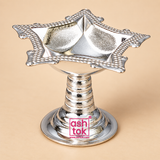 German Silver Diya shown from a three-quarter angle on a warm beige background, highlighting the polished silver-tone metal finish, sturdy circular pedestal base, ringed center stem and the decorative square floral diya bowl with pointed corners and beaded border detailing, captured in soft studio lighting to display the shape, reflective finish and elegant craftsmanship of the diya for use in puja rituals, festive occasions, pooja room decor and traditional gifting.