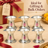 Bulk gifting presentation image showing multiple matching German Silver Diyas arranged neatly on a beige background with elegant festive gift styling, ribbon details and a premium gift box, highlighting their polished silver finish, shallow bowl design, wick-holder extension, tapered conical shape and broad circular bases, making the image suitable for showcasing the product as a return gift, festive gift, pooja favor, wedding favor, housewarming gift or bulk order item for religious and traditional gifting