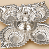 Close-up detail of a German Silver Pooja Diya showing the raised flower shaped central holder, cylindrical wick support, polished reflective silver surface, embossed floral detailing and surrounding decorative diya cups with sculpted ridges and ornamental borders, providing a clear view of the craftsmanship and traditional design for pooja, Diwali, festive decoration and gifting.