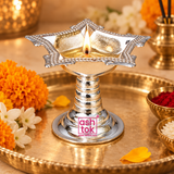 Lifestyle image of a German Silver Diya being used in a traditional puja setting, placed on a decorative brass plate with a lit flame in the center, surrounded by marigold flowers, jasmine flowers and small ritual bowls, showing the diya’s polished silver finish, square floral bowl and pedestal base in a realistic devotional environment, suitable for representing daily worship, festive rituals, pooja room ambiance, Diwali decor and traditional spiritual use without any idol present.