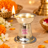 Lifestyle image of a German Silver Diya in use with a lit cotton wick and flame, placed on a warm beige-toned festive surface and surrounded by flowers and traditional decor elements, showing the diya’s polished silver finish, shallow oil bowl, side wick-holder extension, conical body and wide base in a devotional setting, ideal for representing daily puja, festive worship, Diwali decor, pooja room rituals, home temple ambiance and traditional spiritual use without including any idol.