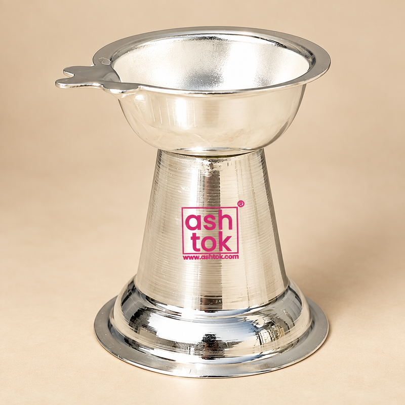 Traditional German Silver Diya with a polished reflective silver finish, featuring a shallow round bowl at the top, a flared rim with a small wick-holder extension on one side, a tapered conical center body and a broad circular flared base, photographed as a clean front-facing product image on a premium beige studio background with soft lighting, showing the full shape clearly for online product display, ideal for daily puja use, festive decor, pooja room placement, return gifting and traditional devotional