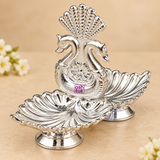 Close-up detail of a German Silver diya showing the elegant double swan centerpiece, floral medallion detail, fan shaped decorative crest and twin leaf shaped diya bowls with a polished silver finish, photographed on a beige background to highlight the fine ornamental craftsmanship and attractive surface texture for pooja decor and premium gifting.