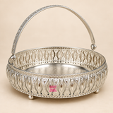 Side angle view of a German Silver Flower Butti decorative basket showing the depth of the round body, elaborate floral cutwork panels, polished silver finish, engraved interior base, raised feet, decorative handle joints and broad arched carrying handle, photographed on a warm beige background to clearly show the three dimensional shape and construction for gifting, serving and festive decorative use.