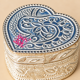 Detailed close-up image of a German Silver Heart-Shaped Kumkum Box focusing on the richly engraved floral and paisley motifs across the heart-shaped lid, decorative dotted border, intricate sidewall patterns and reflective German silver finish, photographed against a premium beige studio background to showcase the fine craftsmanship, ornamental texture, metal detailing and traditional decorative styling of the compact kumkum storage box.