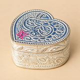 German Silver Heart-Shaped Kumkum Box shown from a three-quarter angle on a premium beige background, highlighting the compact heart-shaped form, engraved floral and paisley lid design, dotted decorative rim, polished metal finish, intricate side patterns and low-profile storage body, presented to clearly show the product shape and craftsmanship for pooja use, festive ceremonies, decorative storage, traditional gifting and return gift purposes.