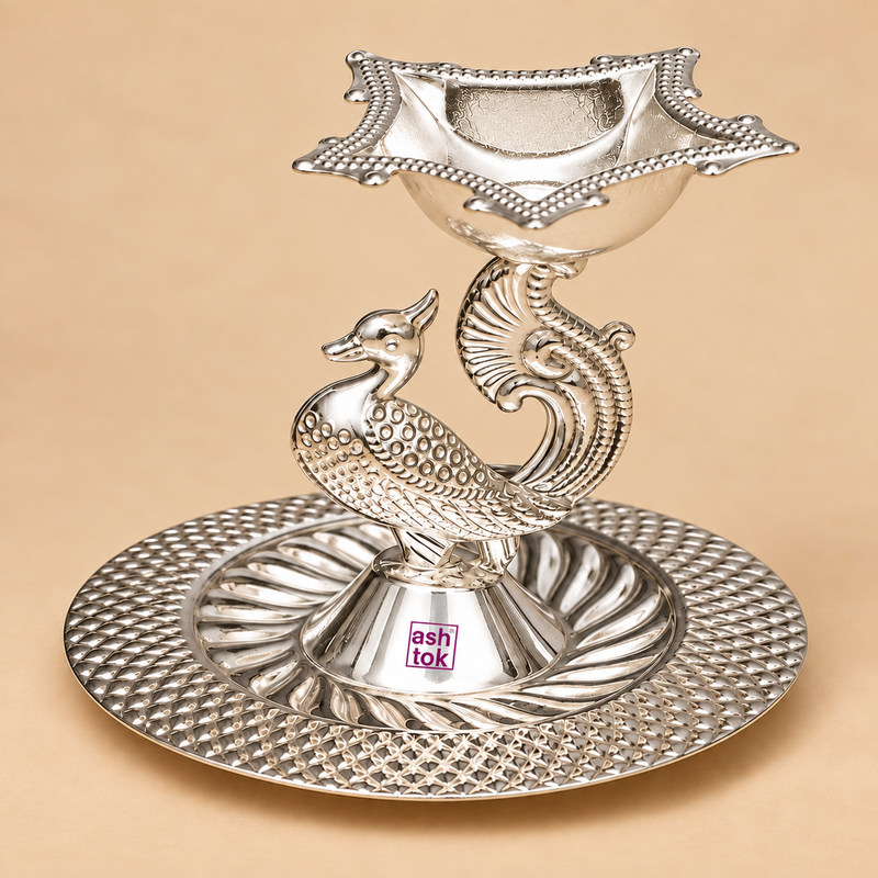 German Silver Pooja Diya with decorative peacock shaped centre stand, raised square oil diya bowl and detailed round floral patterned base, finished in a bright polished silver colour and photographed from the front angle on a warm beige studio background, suitable for daily pooja, home mandir decoration, Diwali celebrations, festive ceremonies, wedding gifts, housewarming gifts and traditional Indian gifting