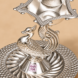 Close-up detail of a German Silver Pooja Diya highlighting the intricately sculpted peacock body, decorative feather patterns, curved tail design, polished silver finish, textured raised diya bowl and embossed circular base, photographed against a beige studio background to clearly show the ornamental craftsmanship and metal detailing for customers considering it for pooja, festive decor and gifting.