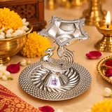 German Silver Peacock Pooja Diya displayed in a traditional festive pooja setting surrounded by marigold flowers, jasmine, rose petals and warm ceremonial lighting, showing the polished silver peacock stand, raised diya bowl and detailed round base as an elegant accessory for daily worship, Diwali decoration, Lakshmi pooja, religious ceremonies and festive home temple arrangements.