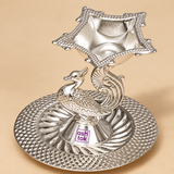 Side view of a decorative German Silver Pooja Diya featuring an intricately designed peacock figure supporting a raised square shaped oil lamp bowl, mounted on a detailed round silver base with embossed leaf and textured patterns, professionally photographed on a beige studio background to show the height, side profile, craftsmanship and traditional design for puja ceremonies and festive home decor.